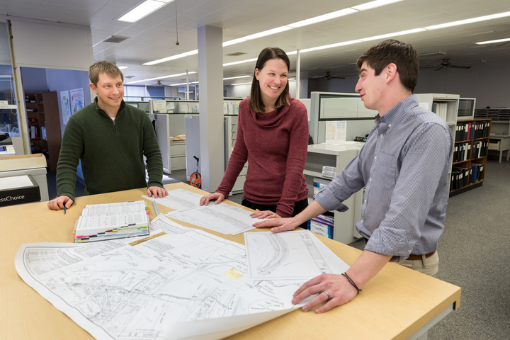two men and one woman stand around an engineering desk looking at plans