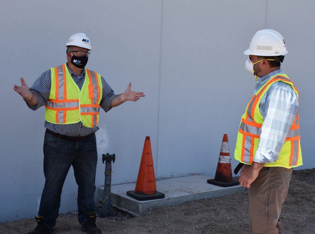 man wearing hard hat, vest, and face mask gesturing to 2nd man